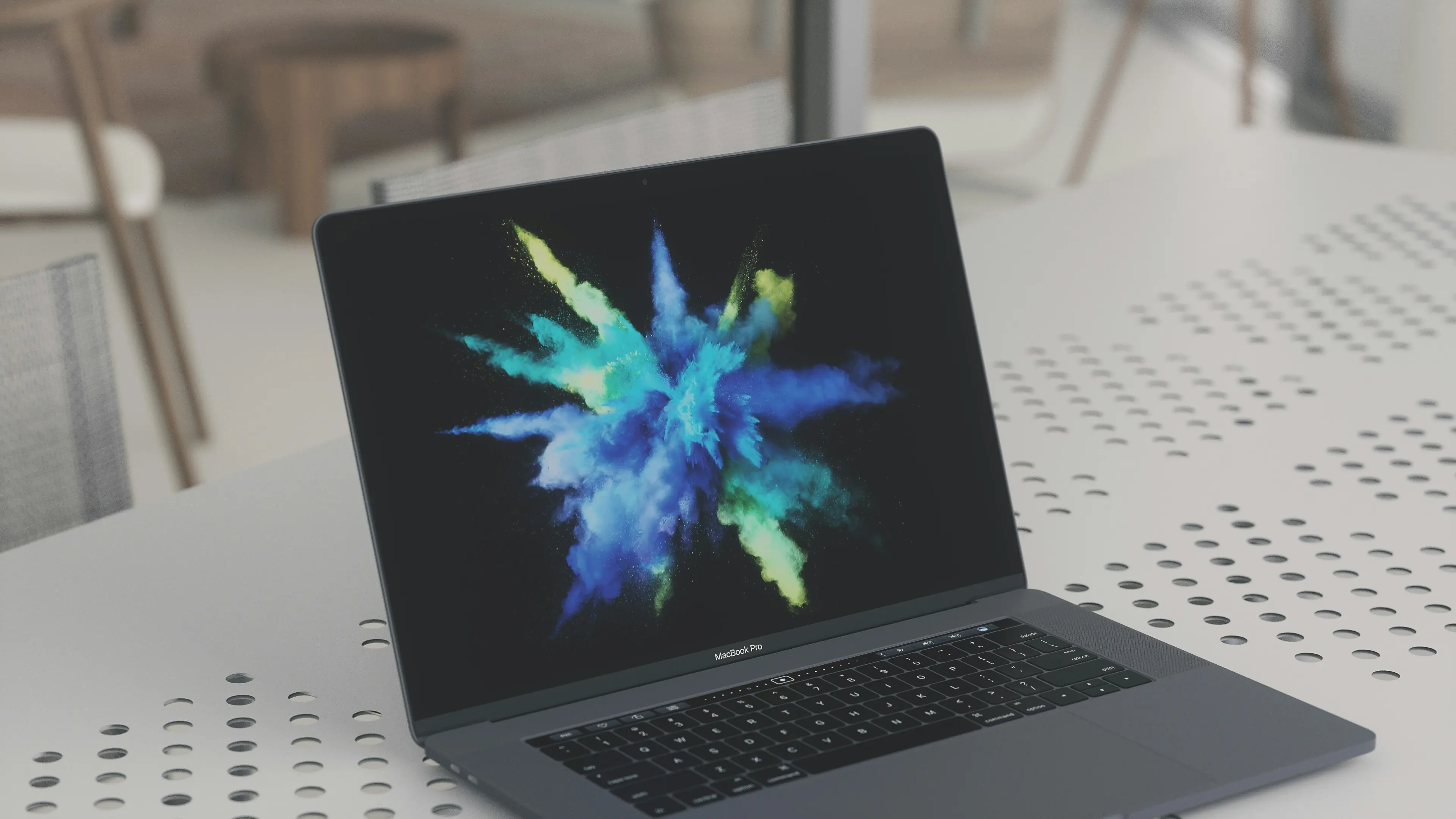 Una imagen de un MacBook Pro con una vibrante pantalla de visualización de colores neón que simbolizan la creatividad y la innovación, características esenciales de los servicios de marketing digital. El portátil está situado en una mesa moderna con un patrón de agujeros en una oficina con decoración minimalista, sugiriendo un entorno de trabajo profesional y contemporáneo.